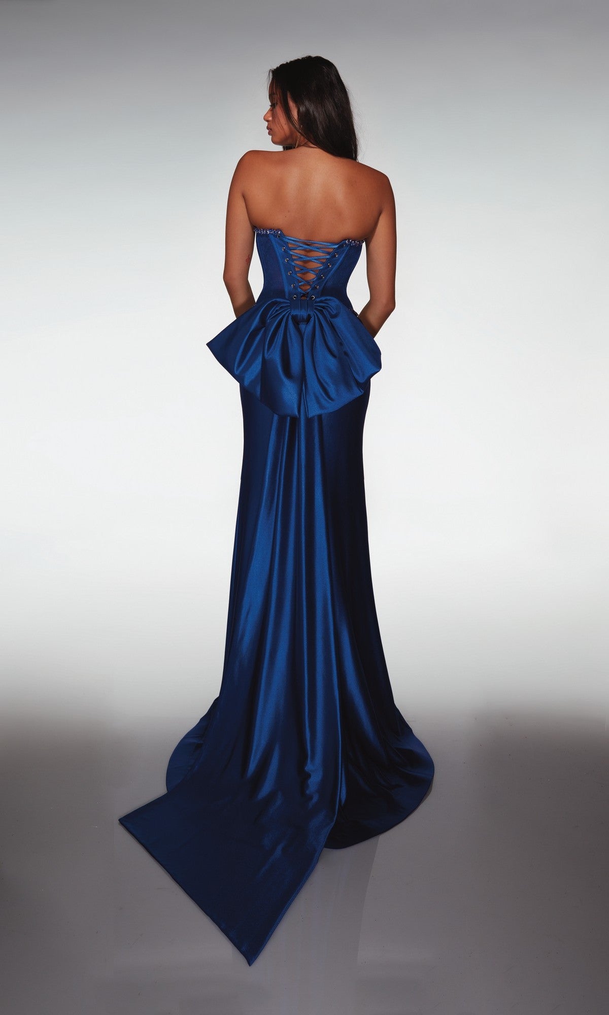 A woman models the Alyce 62089 long prom dress, featuring a strapless fitted lace corset bodice, floor-length royal blue skirt, lace-up back, and large bow, standing with her back to the camera.